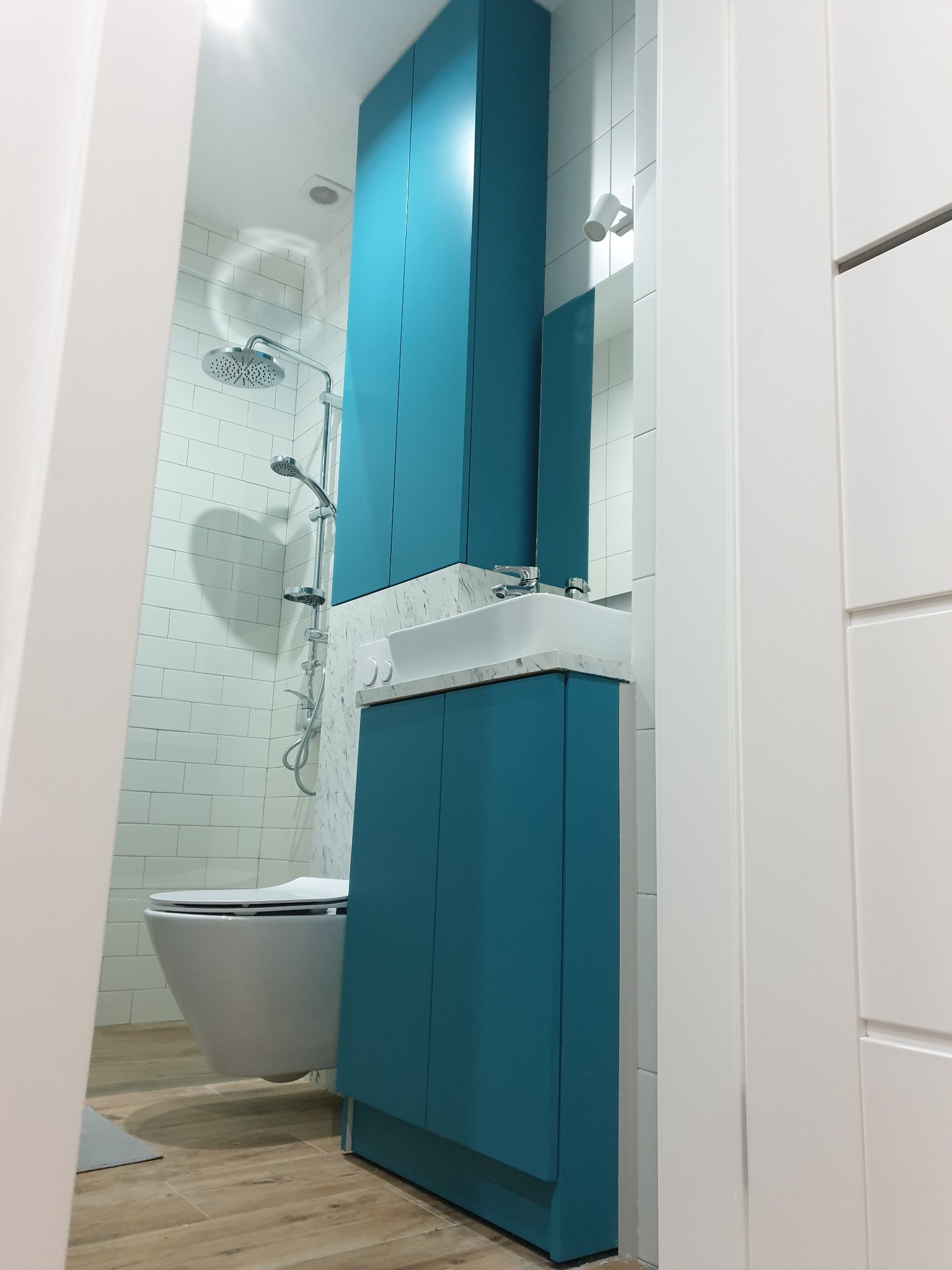 Narożne ujęcie łazienki z turkusową zabudową szafek pod umywalką i wysoką szafą, białą ceramiką sanitarną i kabiną prysznicową wykończoną białymi płytkami.