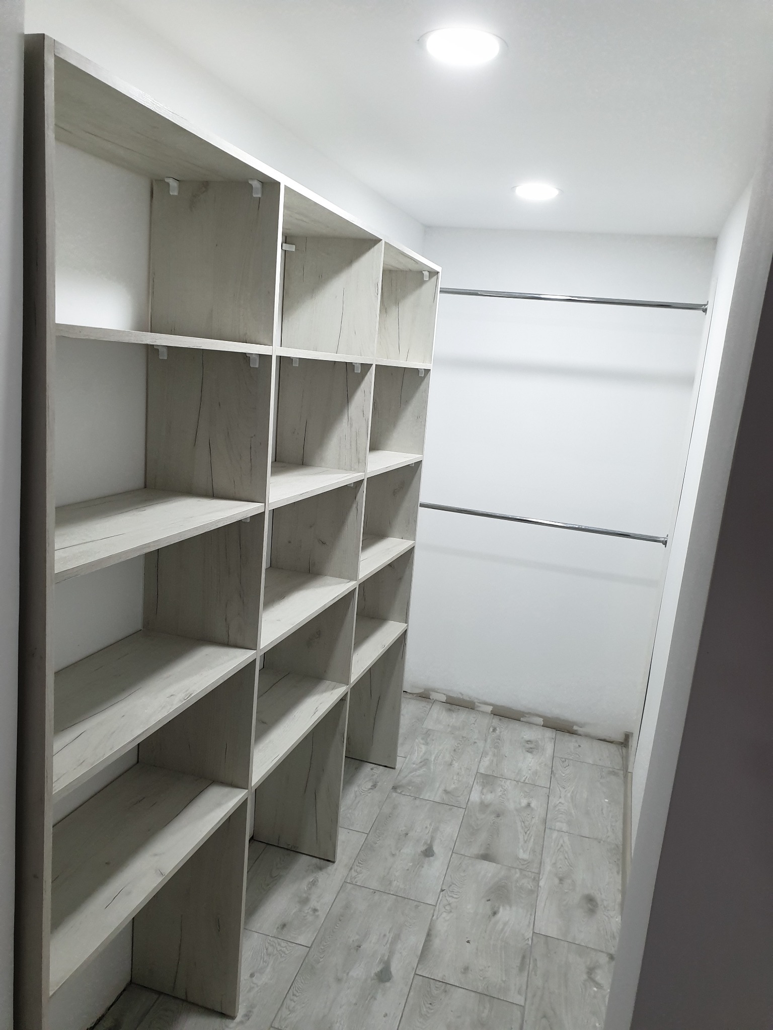 Wnętrze garderoby z półkami z płyty drewnopodobnej w odcieniach szarości i dwoma drążkami na ubrania, oświetlone punktowymi lampami sufitowymi, z podłogą wyłożoną panelami imitującymi drewno.