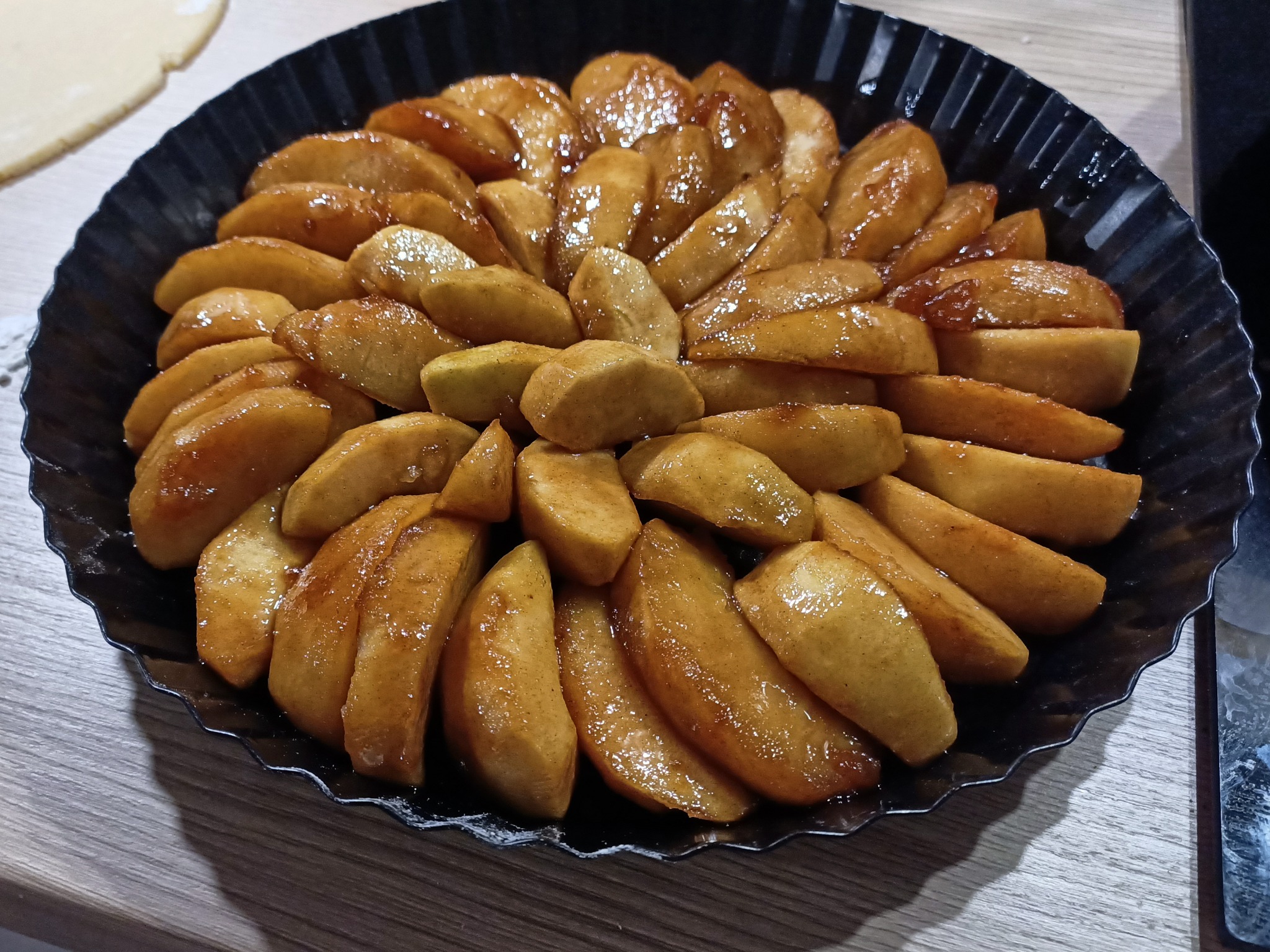 Tarta jabłkowa z błyszczącymi plasterkami jabłek ułożonymi w koncentryczne kręgi na ciemnej, karbowanej formie.