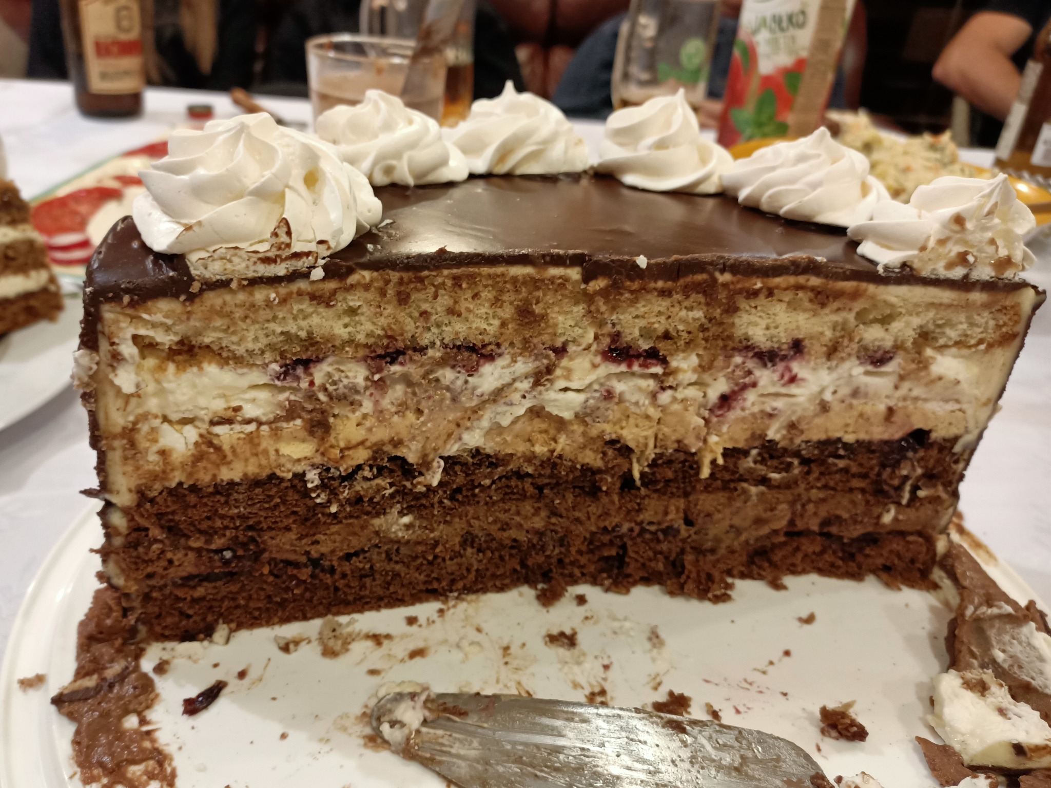 Kawałek tortu z czekoladowym spodem, kremem, warstwą dżemu i polewą czekoladową, udekorowany bitą śmietaną, leżący na talerzu obok srebrnego noża.