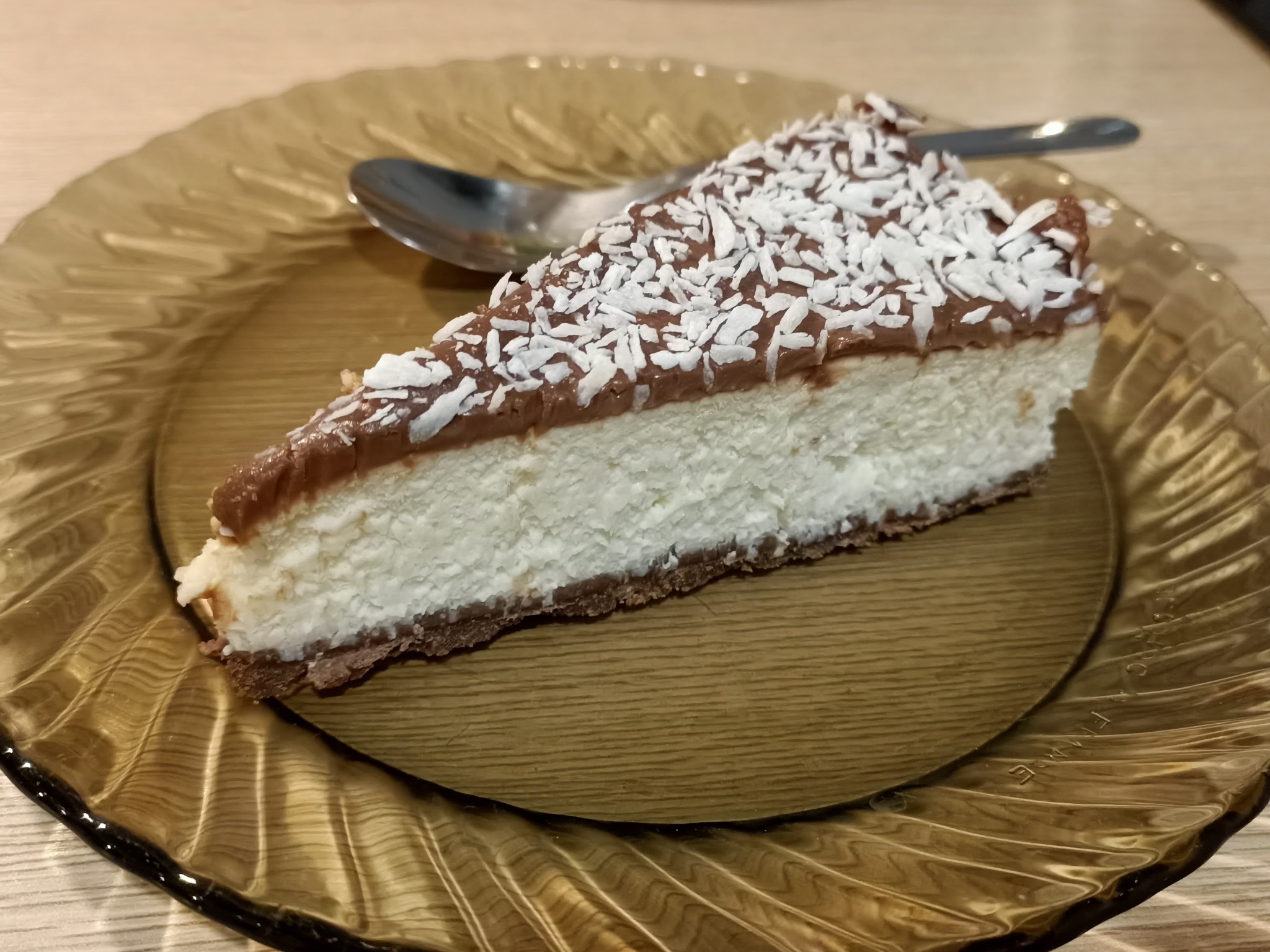 Kawałek sernika z polewą czekoladową i wiórkami kokosowymi na talerzu z przezroczystego, tłoczonego szkła w odcieniach bursztynu, obok łyżeczka.