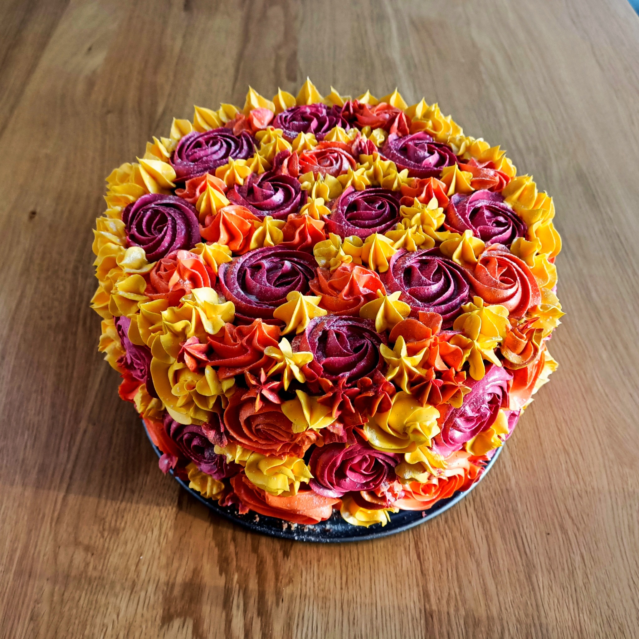 Okrągły tort udekorowany kremowymi różami w odcieniach żółci, pomarańczy i burgundu, umieszczony na drewnianym stole.