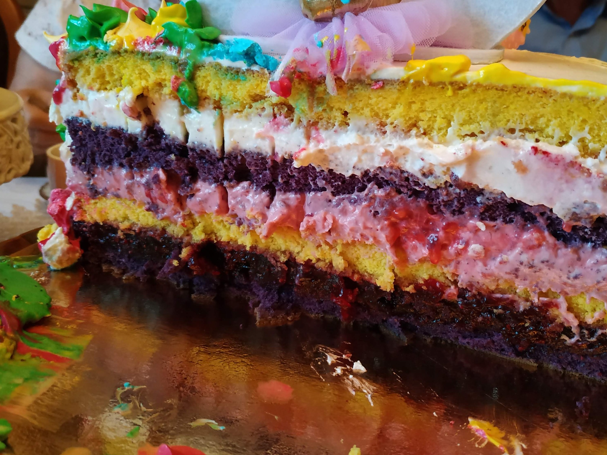 Przekrój wielobarwnego tortu z kremem, widoczne warstwy ciasta w odcieniach żółci i fioletu, dekoracje z kolorowych kremów i tiulu, odbicie na błyszczącej powierzchni.