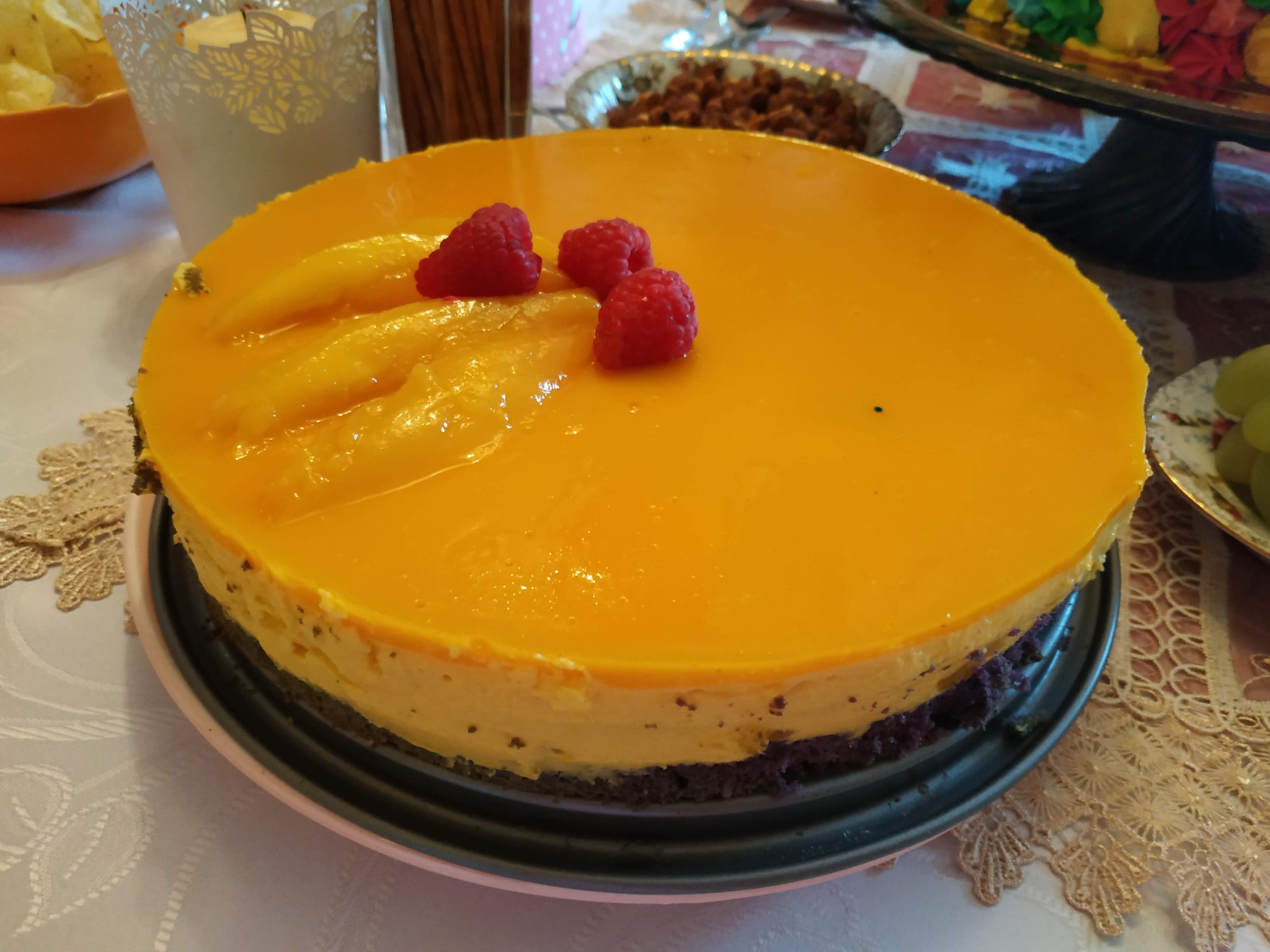Okrągły tort z gładką, pomarańczową polewą, udekorowany kawałkami mango i trzema malinami, na tle świątecznego stołu z innymi przekąskami.