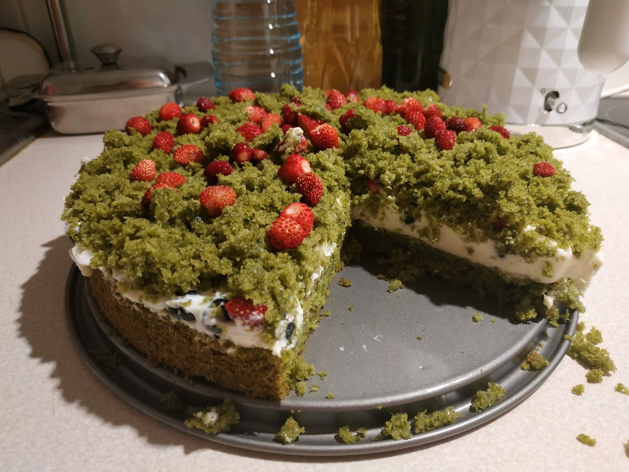 Okrągły tort z zieloną kruszonką i świeżymi poziomkami, z wyciętym kawałkiem, ukazujący kremowe wnętrze z borówkami, umieszczony na metalowej tacy.