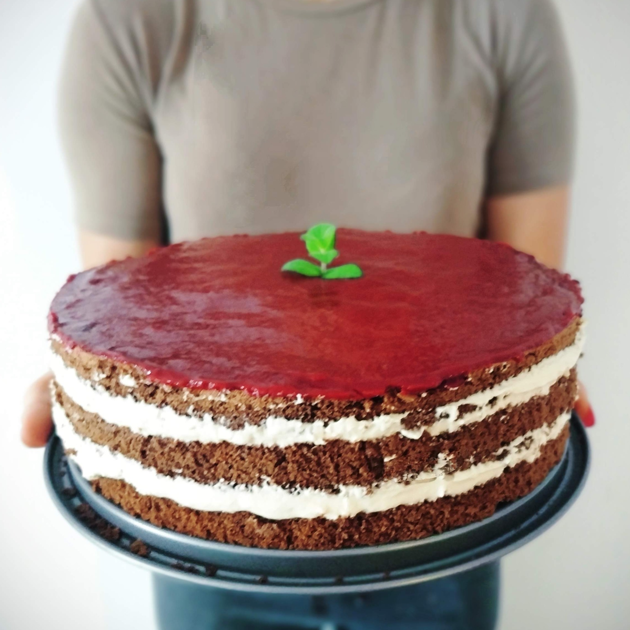 Trójwarstwowy tort czekoladowy z kremem i wiśniową glazurą, udekorowany listkami mięty, prezentowany na blasze.