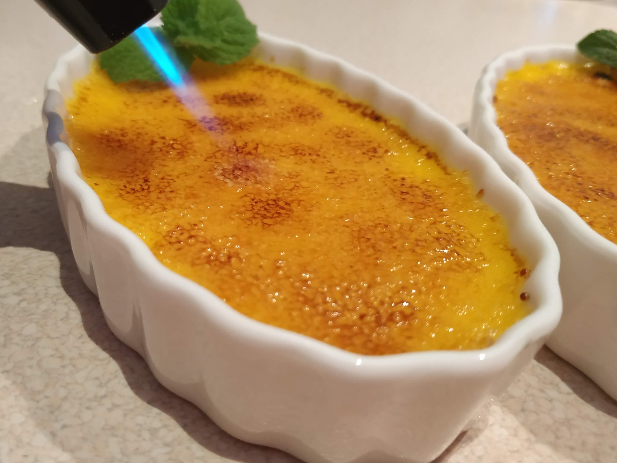 Krem brûlée karmelizowany palnikiem gazowym, zdobiony listkiem mięty w białej ceramicznej miseczce.