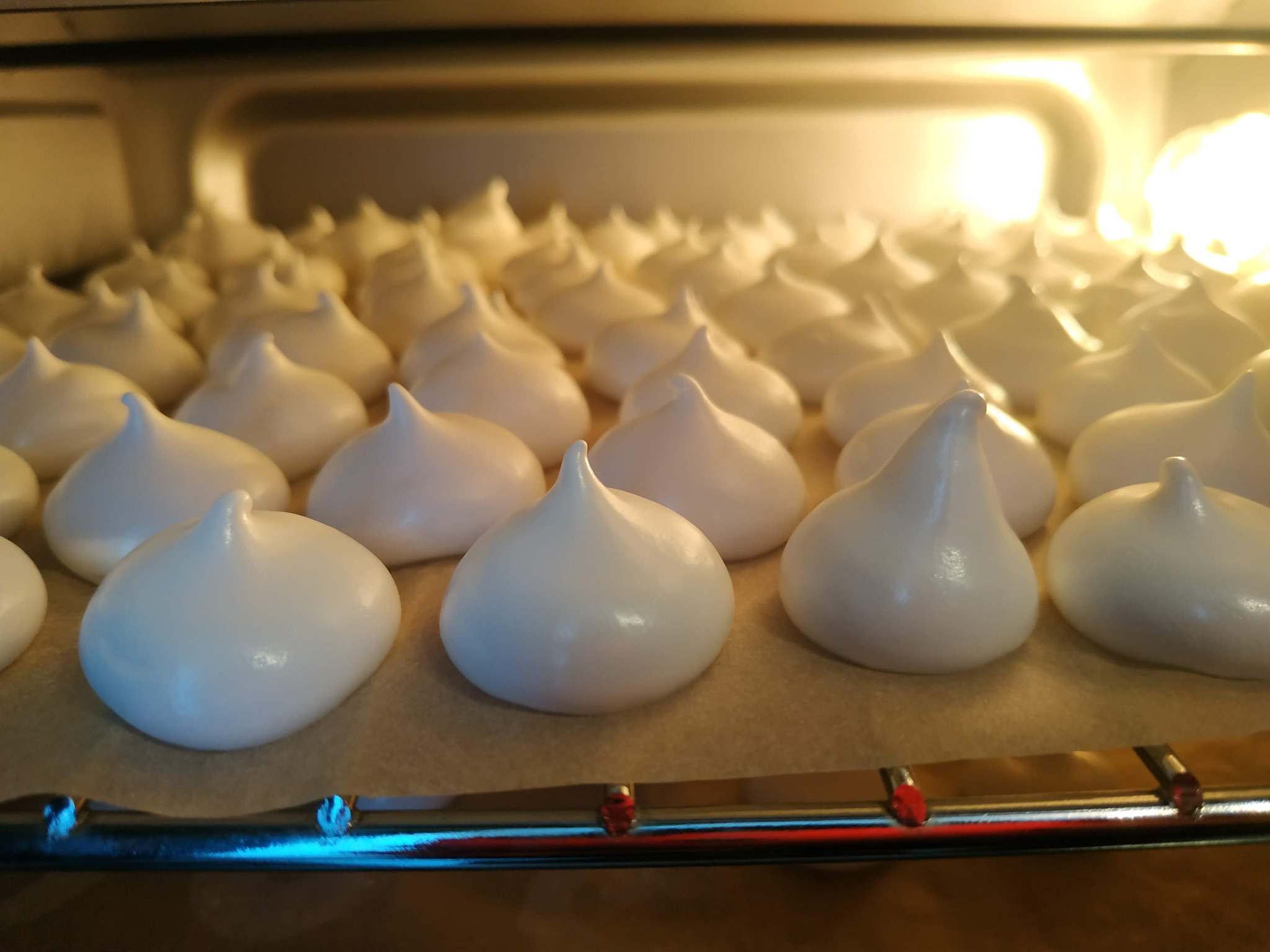 Białe bezy na blasze w piekarniku, oświetlone ciepłym światłem, czekające na upieczenie.