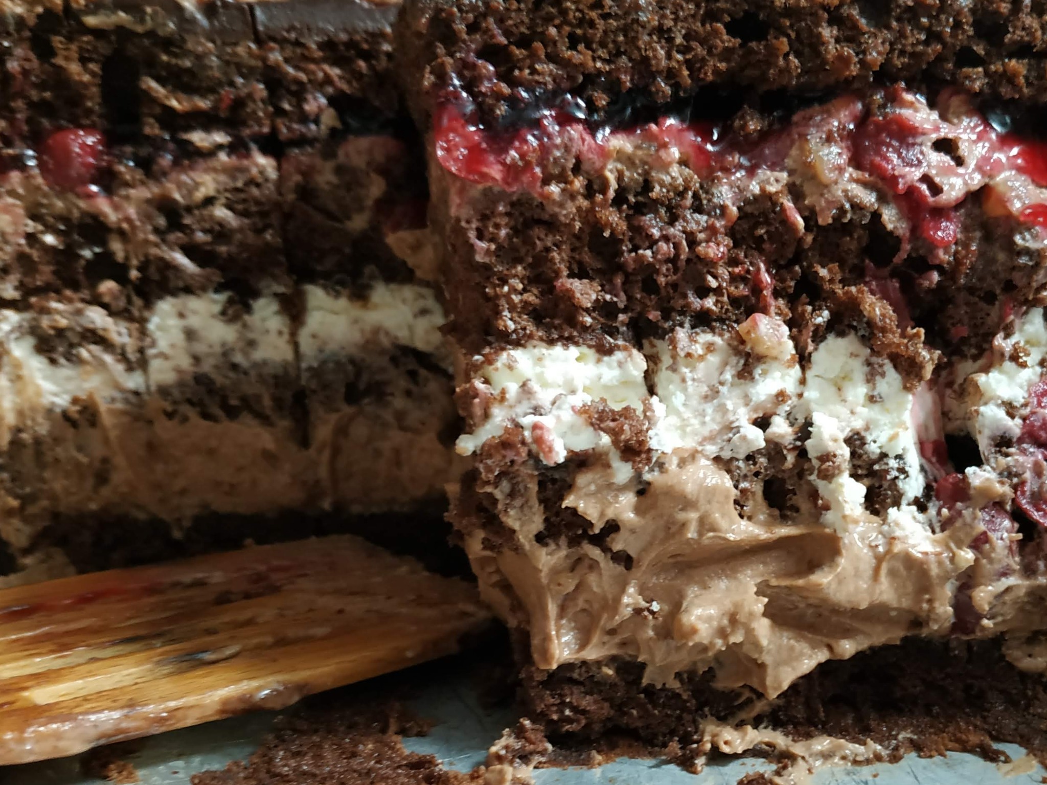 Przekrój wilgotnego tortu czekoladowego z wiśniami, kremem czekoladowym i bitą śmietaną, widoczny drewniany łopatka.
