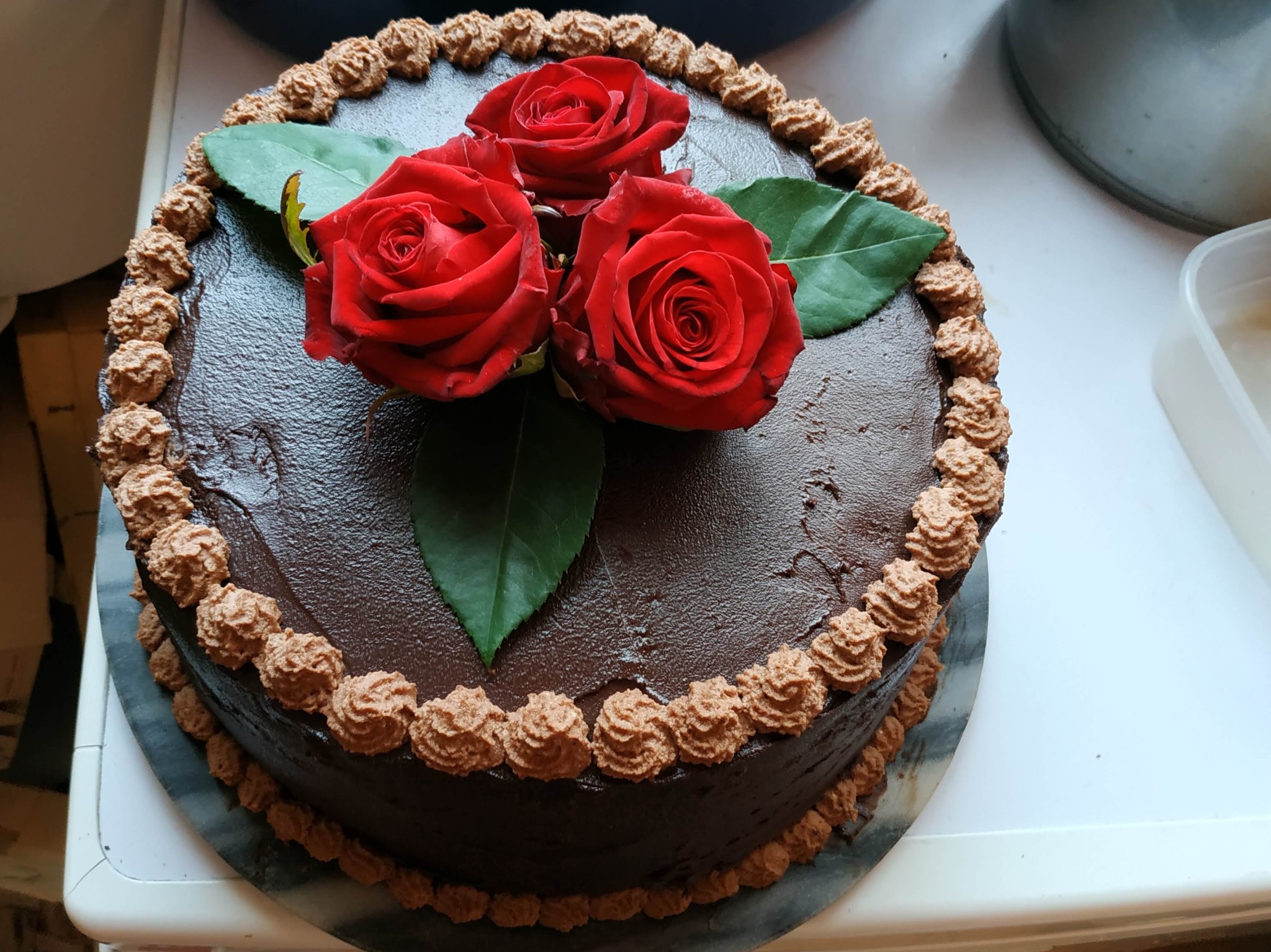 Ciemny tort czekoladowy udekorowany trzema czerwonymi różami i kremowymi rozetkami na obrzeżach.