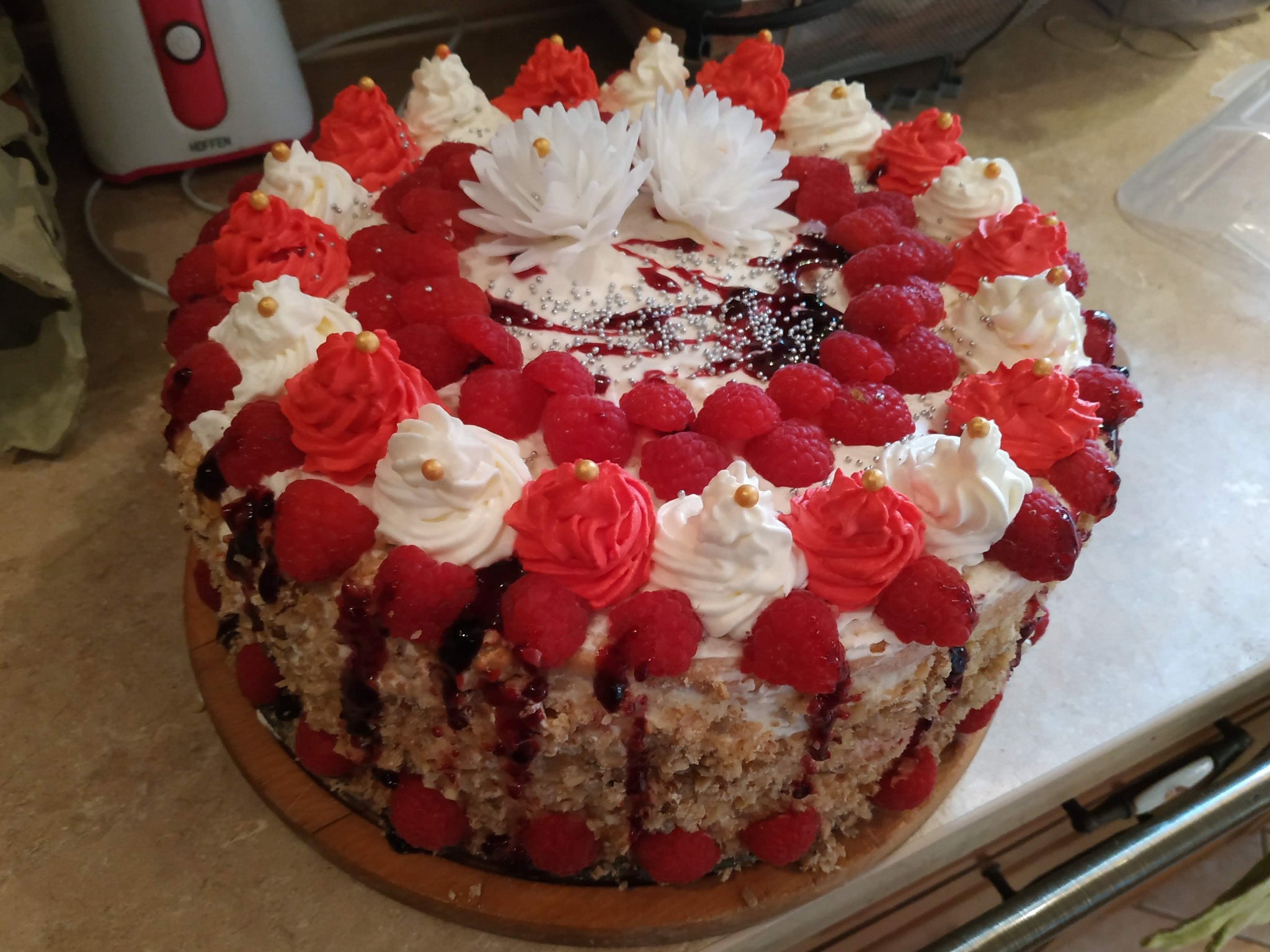 Tort z malinami, kremem i dekoracjami kwiatowymi na drewnianej podstawie, z widocznymi kroplami syropu i srebrnymi kuleczkami.