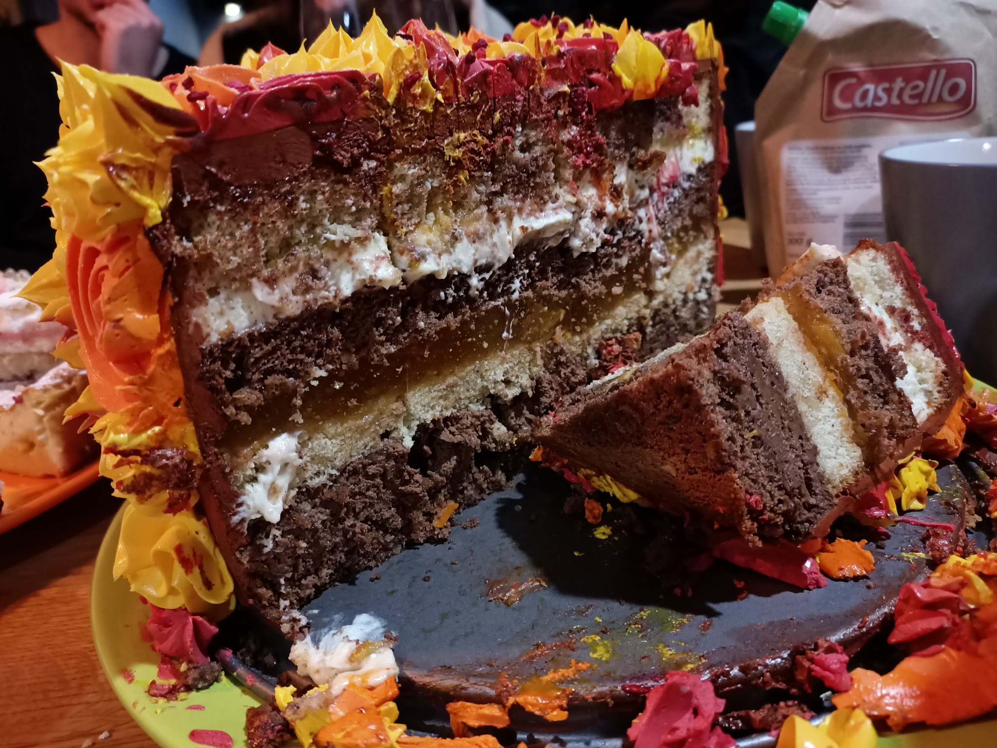 Kawałek warstwowego tortu z kremem w odcieniach żółci i czerwieni, z widocznymi warstwami ciasta czekoladowego, biszkoptu i kremu, leżący na talerzu z resztkami kremu i okruchami, w tle karton...