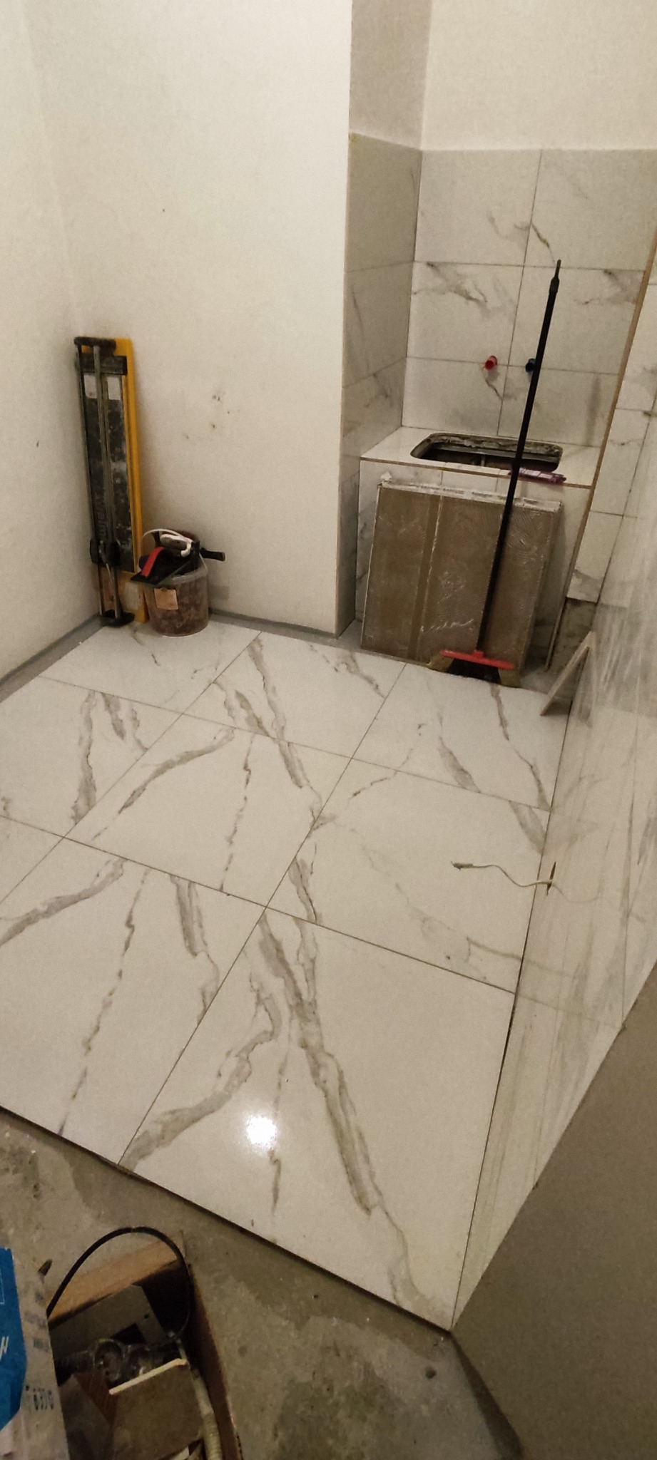 Wykończona podłoga w łazience z białych płytek imitujących marmur, widoczne narzędzia glazurnicze i fragment niezakończonej zabudowy umywalki.