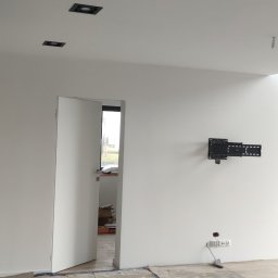 Tom-Bud - Pokój w trakcie remontu: ściany świeżo pomalowane na biało, widoczne czarne oprawy oświetleniowe sufitowe, uchwyt do telewizora na ścianie, podłoga przygotowana do położenia nowej nawierzchni...