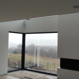 Tom-Bud - Wnętrze w trakcie prac wykończeniowych: widok na duże okno narożne z widokiem na zewnątrz, fragment kominka, podłoga częściowo zabezpieczona folią. Widoczne punkty oświetleniowe w suficie.