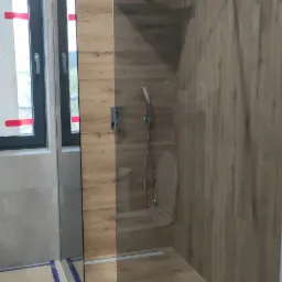 Nowoczesna kabina prysznicowa walk-in z drewnopodobnymi płytkami ściennymi i odpływem liniowym, widoczne zabezpieczenia folią malarską na podłodze.