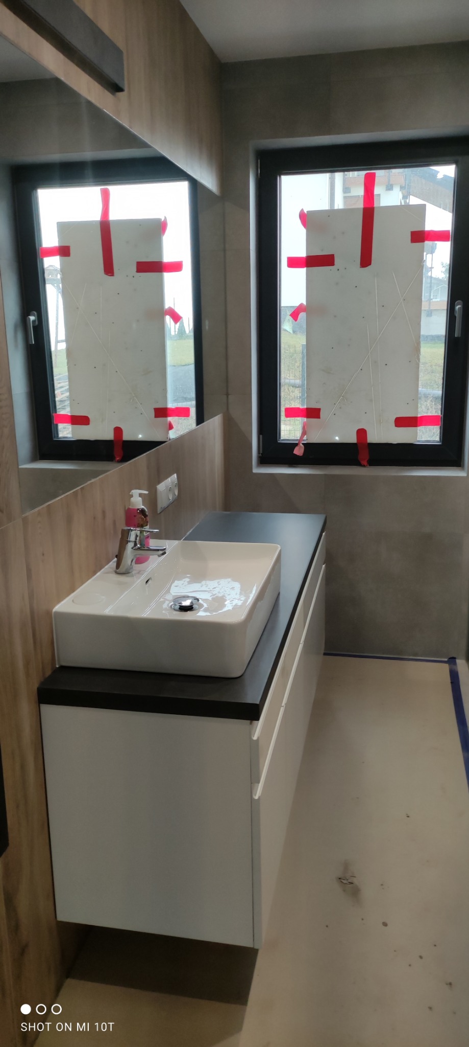 Nowoczesna łazienka w trakcie remontu: biała umywalka nablatowa z armaturą, szare płytki ścienne, okna zabezpieczone folią ochronną z czerwoną taśmą, widoczne ślady prac na podłodze.