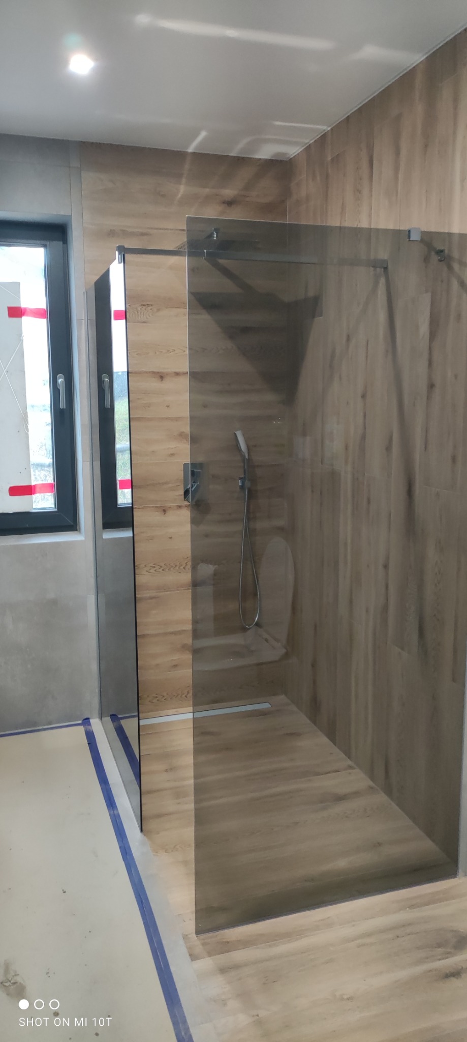 Nowoczesna kabina prysznicowa walk-in z drewnopodobnymi płytkami ściennymi i odpływem liniowym, widoczne zabezpieczenia folią malarską na podłodze.
