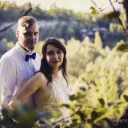 Para zakochanych w plenerze, mężczyzna w białej koszuli i granatowej muszce obejmuje kobietę w koronkowej sukience na tle zieleni i drzew