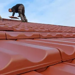 Montaż brązowych dachówek ceramicznych na dachu domu przez dekarzy, widok z dołu pod kątem, jasne niebo w tle.