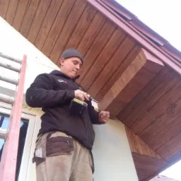 Monter w czapce, z wiertarką, mocuje drewnopodobną podbitkę dachową na zewnątrz budynku, widoczna drabina i fragment dachu z dachówki ceramicznej.