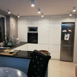 Nowoczesna kuchnia z białymi, lakierowanymi szafkami, czarnym blatem i sprzętem AGD w zabudowie, oświetlona punktowymi lampami sufitowymi; na blacie deska z masłem i miską.