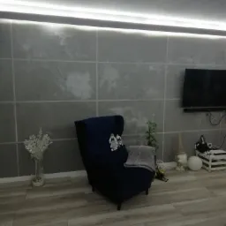 Szary salon z ciemnoniebieskim fotelem typu uszak, dekoracyjną ścianą imitującą płytki i podświetleniem LED pod sufitem.