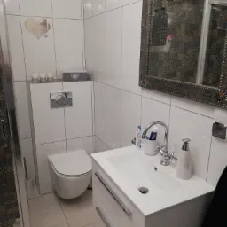 Nowoczesna łazienka z białymi płytkami, wiszącą toaletą, białą szafką pod umywalką i ozdobnym lustrem z wzorzystym odbiciem płytek prysznicowych.