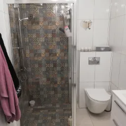 Wykończona łazienka z kabiną prysznicową wyłożoną mozaiką w odcieniach brązu i szarości, wiszącą toaletą i białymi płytkami ściennymi.