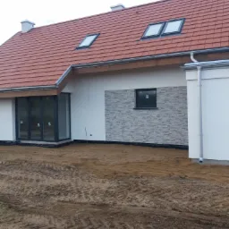 Nowoczesny dom jednorodzinny z czerwoną dachówką i oknami dachowymi, elewacja częściowo wykończona szarym kamieniem dekoracyjnym, teren wokół domu w trakcie wyrównywania.