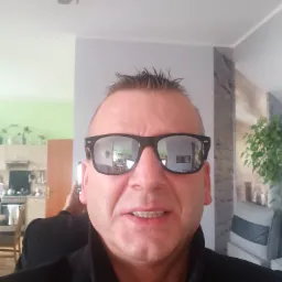 Autoportret mężczyzny w okularach przeciwsłonecznych, w których odbija się wnętrze mieszkania z widoczną kuchnią i dekoracyjnym panelem ściennym.