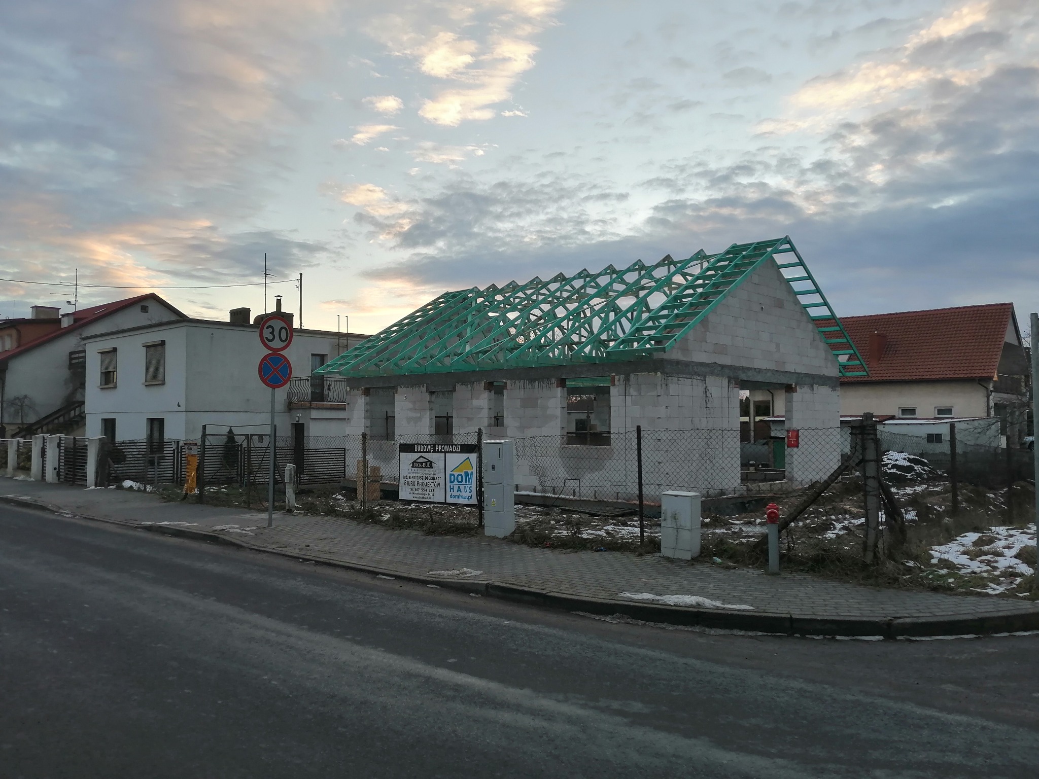 Surowy stan budynku z pustaków z zieloną konstrukcją dachu, widok z ulicy, znak drogowy 30 km/h w tle.