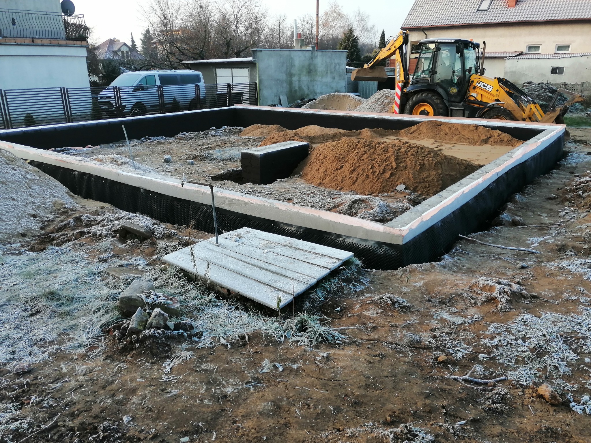 Wykopany i przygotowany fundament pod budynek z zaizolowanymi ścianami i stertami piasku w środku, na zewnątrz ziemia pokryta szronem, w tle koparka JCB i zabudowania.