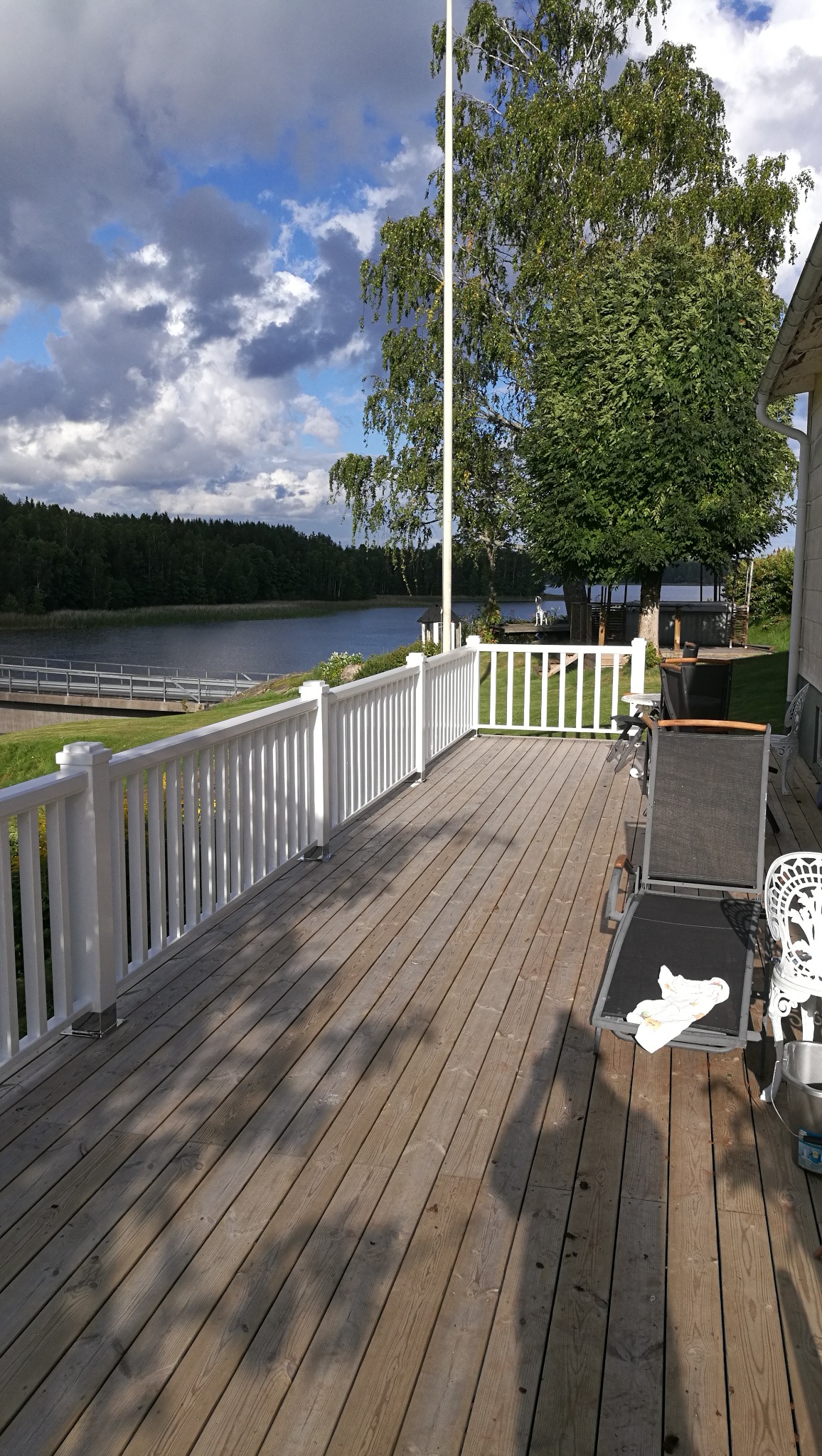 Drewniany taras z białymi balustradami, widok na jezioro i las, częściowo zacieniony, z leżakiem i krzesłem.