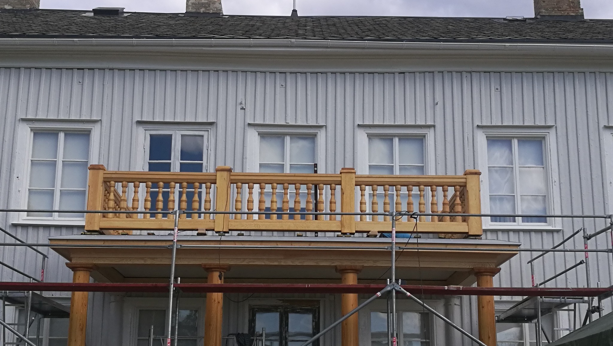 Drewniany, odrestaurowany balkon z balustradą w stylu retro, zamontowany na tle szarej, drewnianej elewacji budynku, widoczny z poziomu rusztowania.