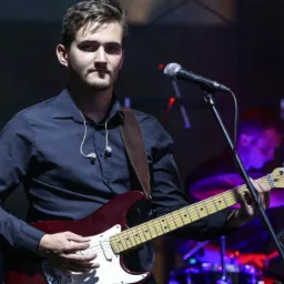 Młody mężczyzna gra na elektrycznej gitarze Fender Stratocaster, ubrany w czarną koszulę z podwiniętymi rękawami, występuje na scenie z mikrofonem i perkusją w tle.