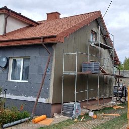 MalBud - Ocieplanie budynku styropianem: ściana szara z widocznymi płytami styropianowymi i rusztowaniem, fragment koparki z numerem telefonu widoczny w prawym dolnym rogu.