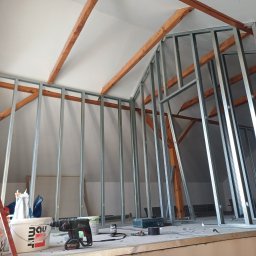 Szkielet ścianki działowej z profili stalowych na poddaszu, widoczne drewniane elementy konstrukcyjne dachu, narzędzia i materiały budowlane w tle. Etap prac wykończeniowych.