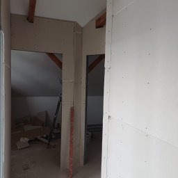 MalBud - Poddasze w trakcie zabudowy płytami gipsowo-kartonowymi, widoczne drewniane belki stropowe i fragment poziomicy. Prace wykończeniowe w surowym stanie.