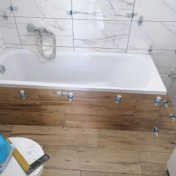 Montaż płytek imitujących drewno na obudowie wanny i podłodze w łazience. Widoczne krzyżyki dystansowe i białe płytki marmurowe na ścianach. Etap prac wykończeniowych.