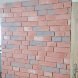 Ściana wykończona dekoracyjną cegłą w odcieniach czerwieni i szarości, przylegająca do drewnianej ościeżnicy drzwiowej z widoczną klamką. Nowoczesny design wnętrza.