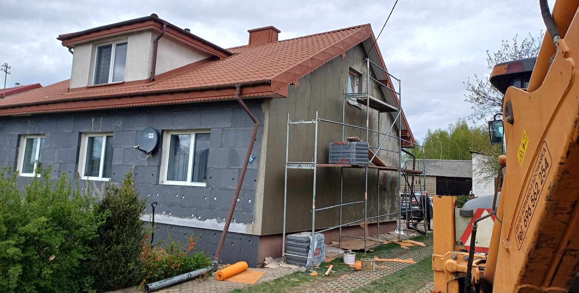 Ocieplanie budynku styropianem: ściana szara z widocznymi płytami styropianowymi i rusztowaniem, fragment koparki z numerem telefonu widoczny w prawym dolnym rogu.