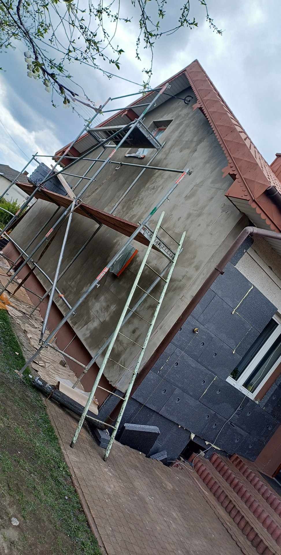 Elewacja domu w trakcie ocieplania: fragment ściany pokryty szarym tynkiem, część z ciemnoszarymi płytami izolacyjnymi, rusztowanie i drabina przy ścianie, widoczne elementy dachu i okna.