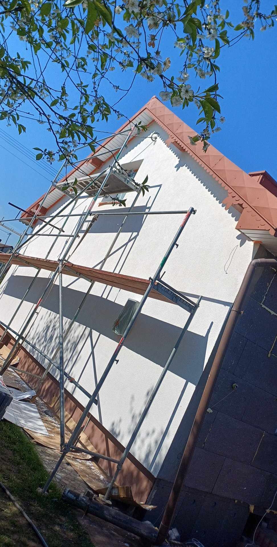 Dom jednorodzinny w trakcie ocieplania, widoczne rusztowanie, fragment elewacji pokryty białą warstwą izolacyjną, częściowo widoczne ocieplenie fundamentów, na pierwszym planie kwitnące gałęzie...