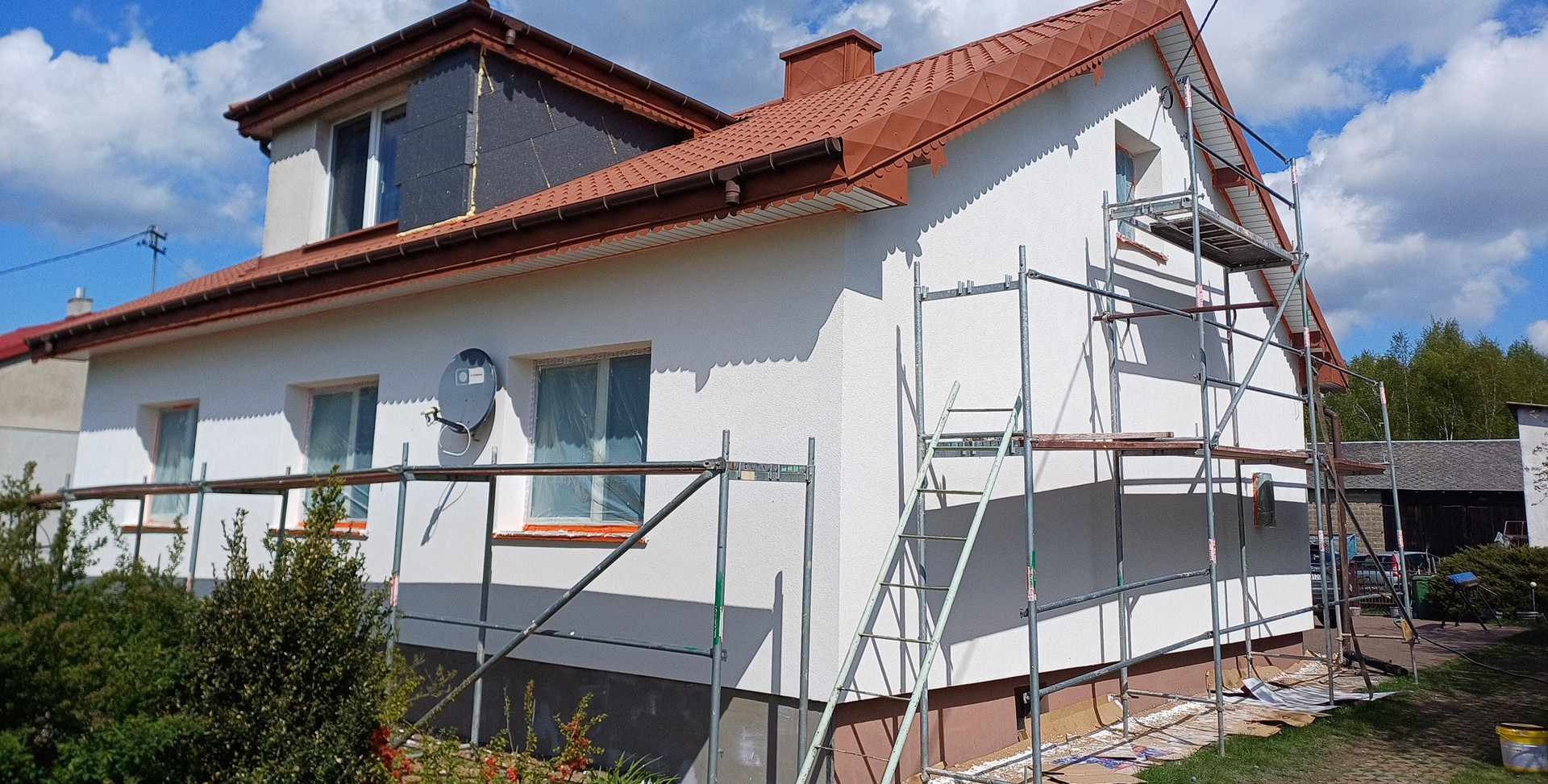 Dom jednorodzinny z nową, białą elewacją, częściowo osłoniętymi oknami i rozstawionym rusztowaniem, w tle błękitne niebo z chmurami.