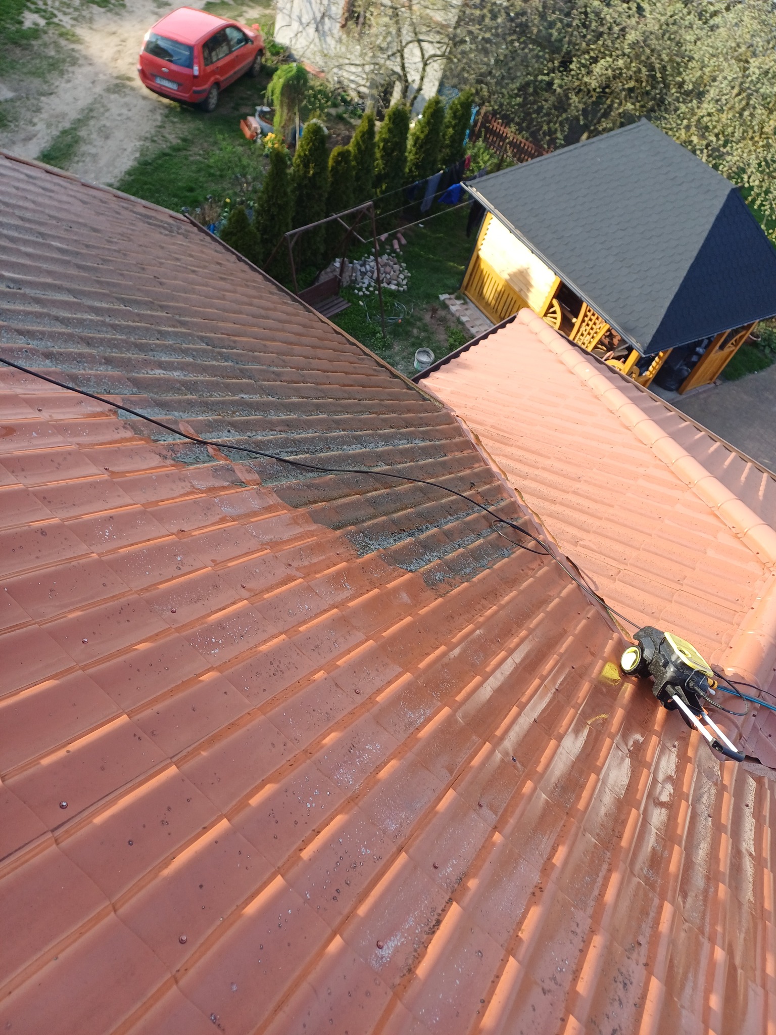 Częściowo wyczyszczony, czerwony dach z dachówki ceramicznej, z widocznym urządzeniem czyszczącym i ogrodem z altaną w tle.