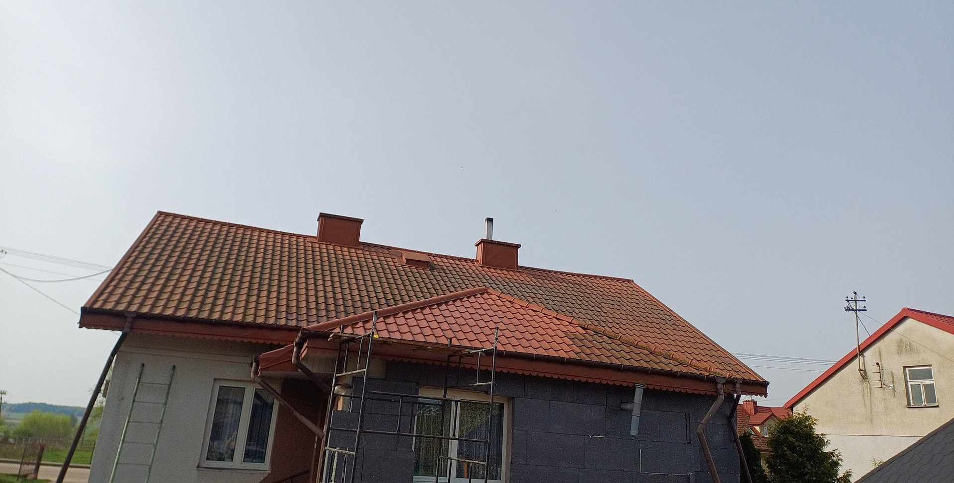 Dom w trakcie remontu elewacji i dachu z widocznymi rusztowaniami, nowa czerwona dachówka i szara elewacja z bloczków betonowych.