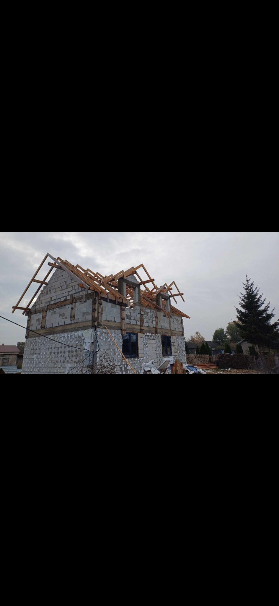 Surowy stan budowy domu jednorodzinnego z pustaków, z widoczną konstrukcją drewnianą dachu, w tle iglaste drzewo i pochmurne niebo.