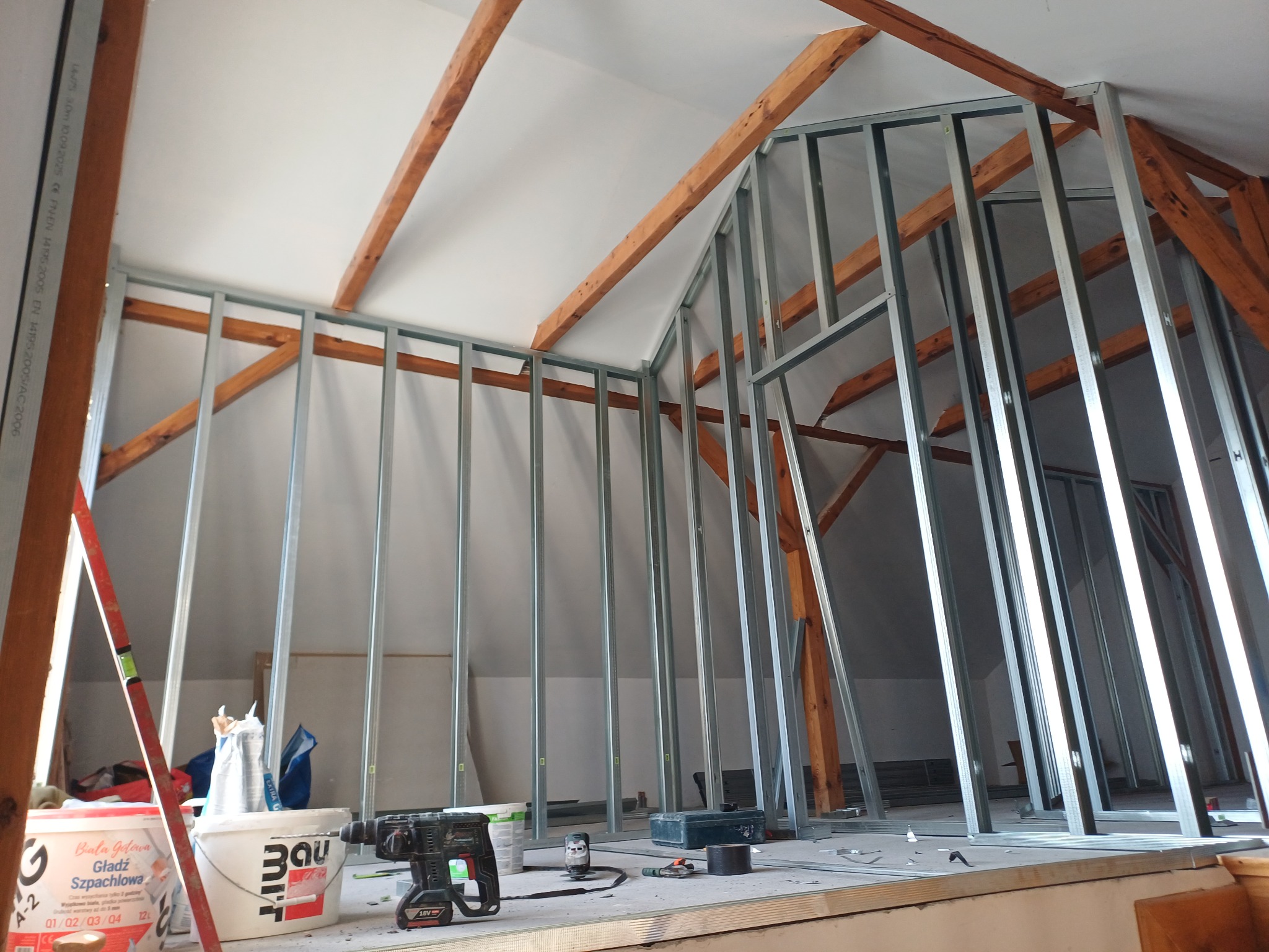 Szkielet ścianki działowej z profili stalowych na poddaszu, widoczne drewniane elementy konstrukcyjne dachu, narzędzia i materiały budowlane w tle. Etap prac wykończeniowych.