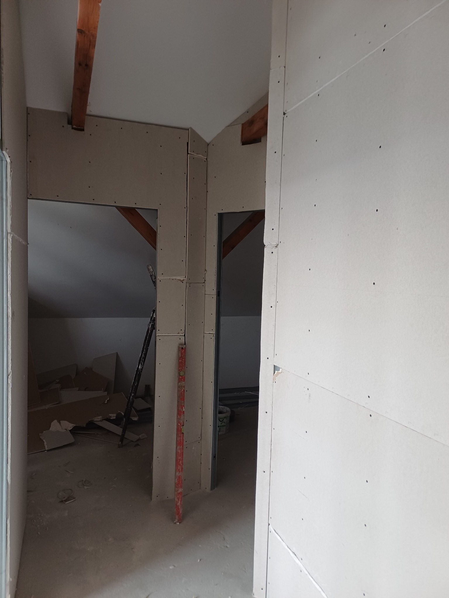 Poddasze w trakcie zabudowy płytami gipsowo-kartonowymi, widoczne drewniane belki stropowe i fragment poziomicy. Prace wykończeniowe w surowym stanie.